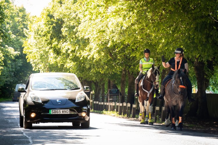 Two horses being ridden down a road and being passed by an electric car