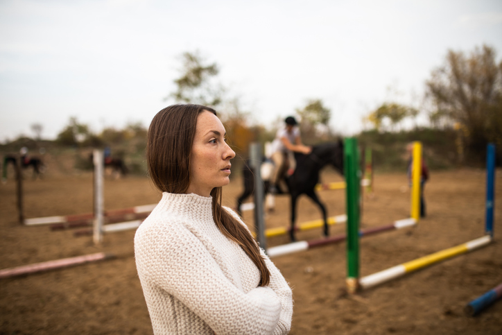Adult female standing in ring with horse and rider negotiating cavaletti in the background
