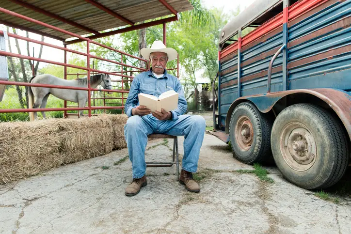 Seated man in cowboy hat reading book with horse and trailer in background