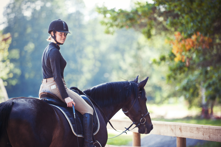 Young pretty cheerful girl resting after horseback riding training on arena. Vibrant colored outdoors horizontal summertime image with vintage filter. View from back