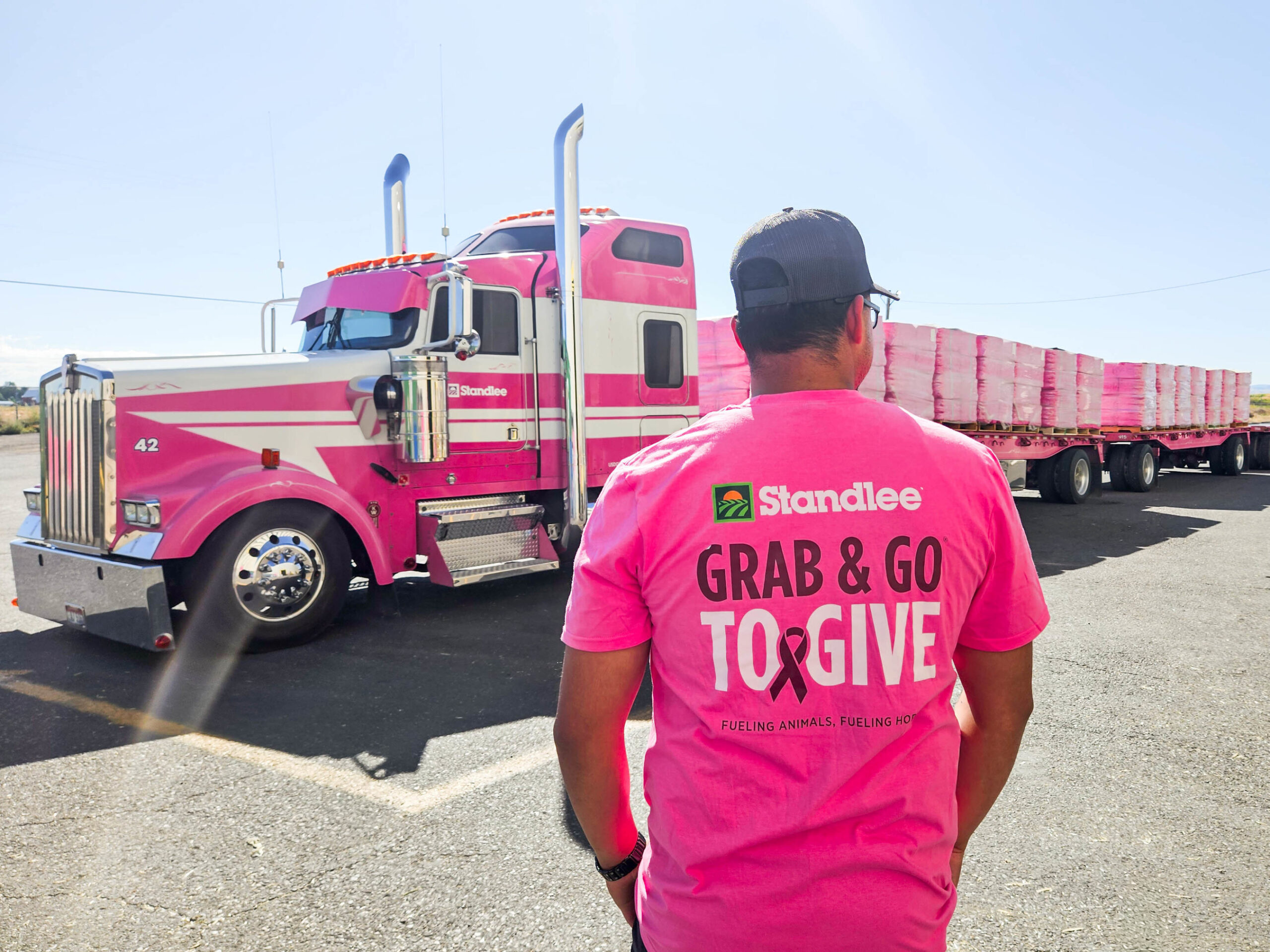 Man wearing pink Standlee "Grab & Go to give" shirt and trucker hat faces away from the camera and toward a pink tractor-trailer