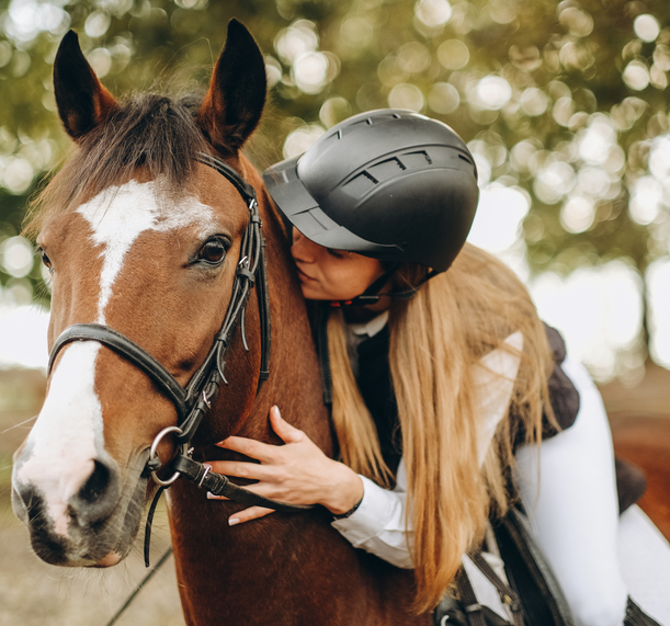 A young rider with long hair and a black helmet leans forward from the saddle and huge her bay horse, who has a white blaze and a bridle